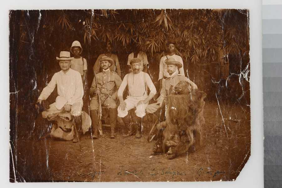 Yellowed photo: Four German colonial officials with hunting weapons and hunted animals in the foreground, sitting, behind them four African young men, likely informants or hunters, standing. © MfN Berlin