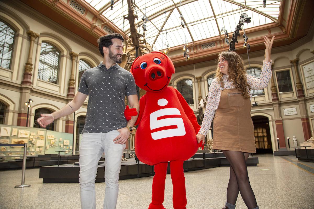 Süßes oder Saurier: Die Moderator:innen und Sparky, Maskottchen der Berliner Sparkasse, im Sauriersaal.