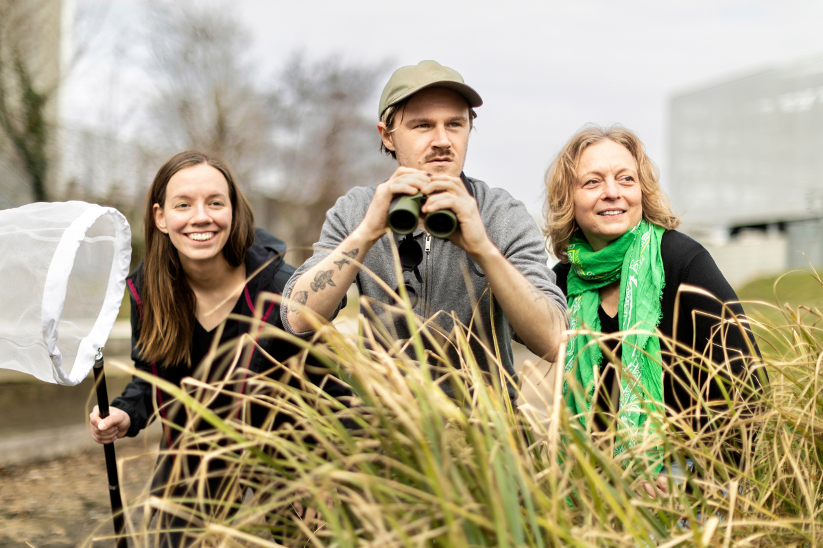Drei Personen im Vordergrund mit einem Ferngläsern und mehreren Pflanzen um das Trio. 