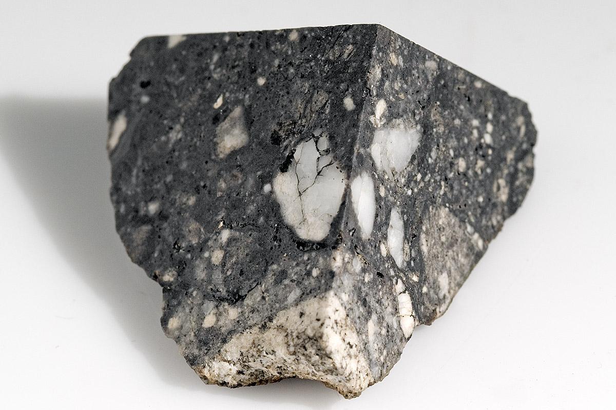 Das Foto zeigt einen Mondmeteoriten. Er besteht aus Bruchstücken unterschiedlicher Minerale und Gesteine (Brekzien). Die farbigkeit variiert zwischen dunkelgrau bis hellgrau und weiß.
