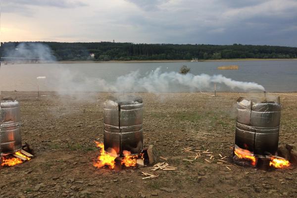 Vor einem See stehen drei Feuerstellen mit Fässern, aus denen Rauch aufsteigt. Die Fässer werden zum Köhlern verwendet.