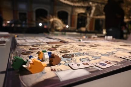 A board game lies spread out on a table against the background of the skeletons in the Dinosaur Hall of the Museum für Naturkunde Berlin